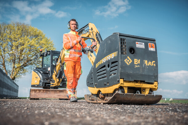 Arbeiter mit kleiner Vibrationswalze auf Baustelle unter blauem Himmel.